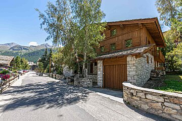 A charming wooden and stone chalet on a sunny village street with mountains in the background, surrounded by trees.