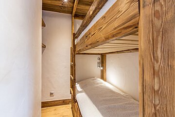 A rustic wooden bunk bed with white textured walls. Spotlights illuminate the cozy lower bunk, covered with a white blanket.