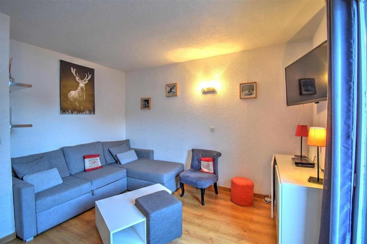 A cozy living room features a gray L-shaped sofa, white coffee table, wall-mounted TV, and deer artwork, with warm lighting and wooden floors.