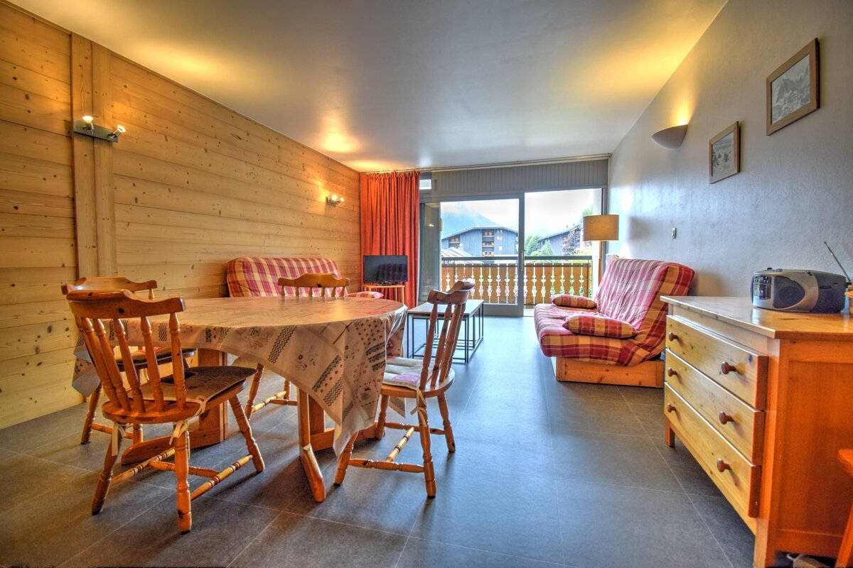 A cozy, rustic living and dining space with wood-paneled walls, plaid furniture, a dining table, dark floor, and a balcony offering mountain views.