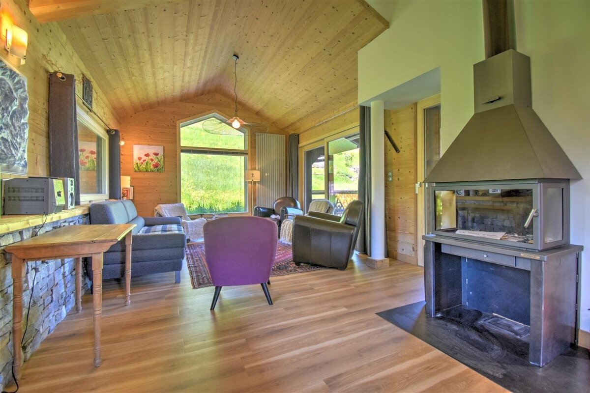 A cozy, wood-paneled living room with a modern fireplace, a purple armchair, and large windows revealing a lush green outdoor landscape.