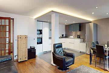 Modern open-plan kitchen, dining, and living area with white cabinets, wood floors, stainless steel appliances, and a black leather armchair.