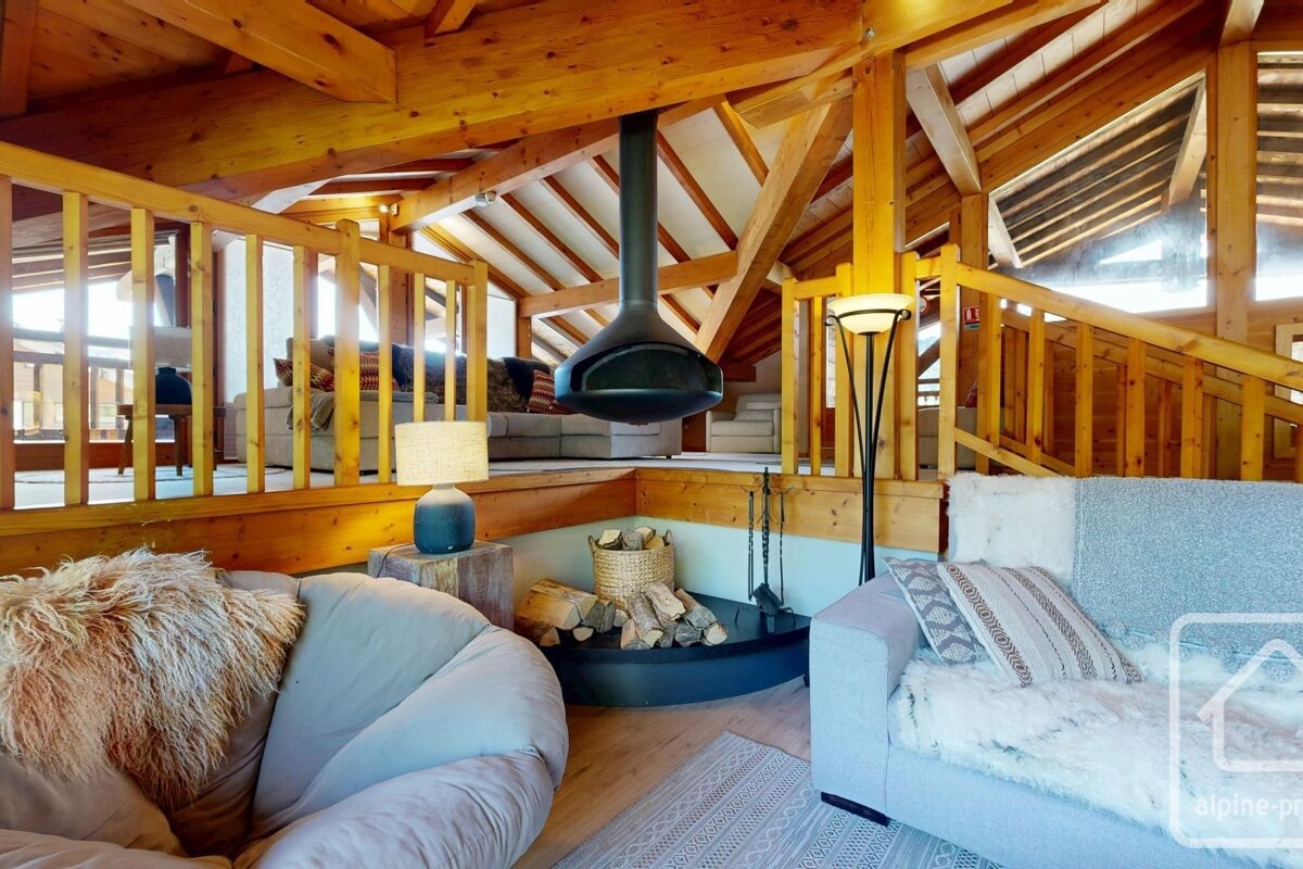 Cozy, multi-level wooden cabin interior with exposed beams, comfortable seating, and a central hanging black fireplace.