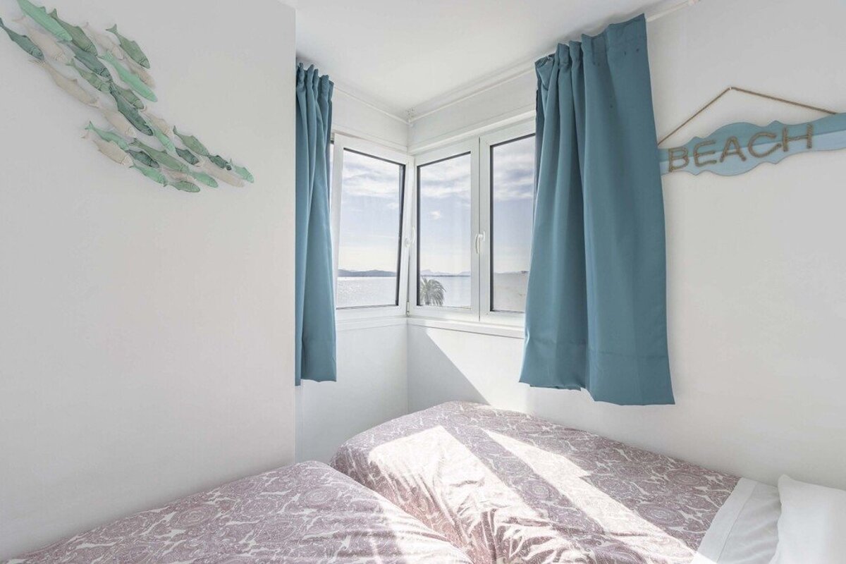 Bright bedroom with two single beds, blue curtains, fish art, beach sign, and a window with sea views.