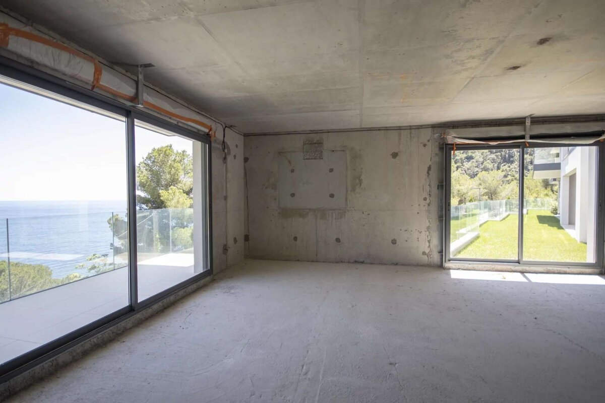 An empty, unfinished concrete room with panoramic windows offering views of the sea and a green garden courtyard.
