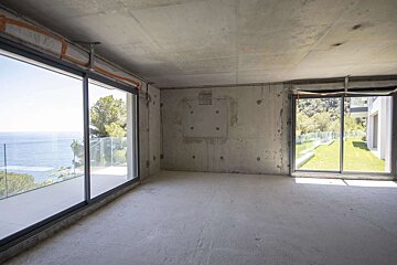 An empty, unfinished concrete room with panoramic windows offering views of the sea and a green garden courtyard.
