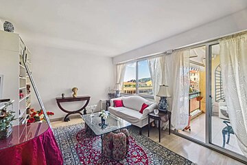 A bright living room with a white sofa, red rug, glass table, and a large window overlooking a view and balcony. Bookshelf with a ladder on the left.