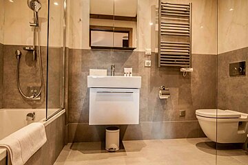 A modern bathroom featuring a tub with shower, sleek white sink, toilet, and a heated towel rack. Walls are tiled in neutral grey and beige.
