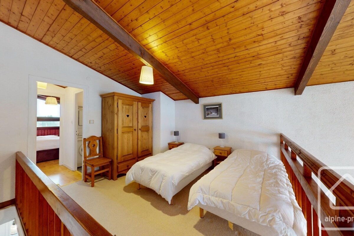 A cozy loft bedroom featuring a wooden vaulted ceiling, two single beds, a wooden wardrobe, and a railing overlooking the floor below.