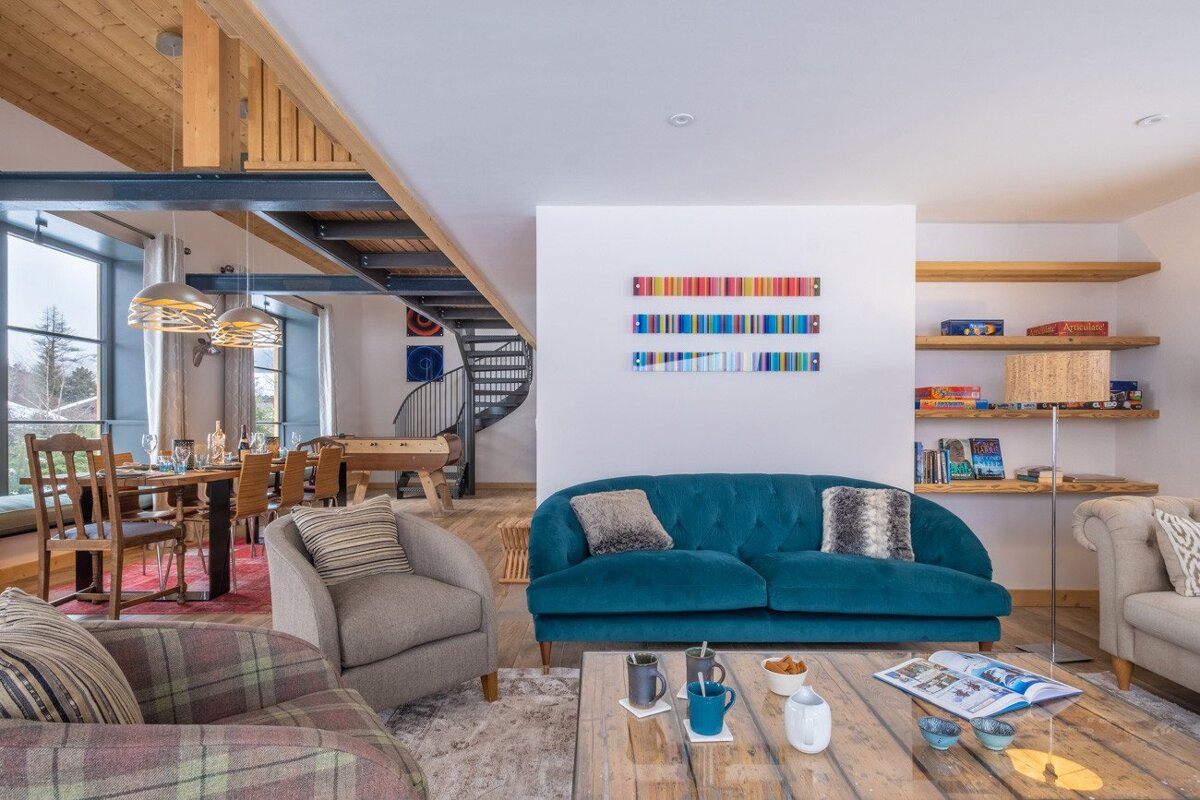 Bright, open-plan living area with a teal sofa, dining space, spiral staircase, and a blend of modern design with rustic wooden beams.