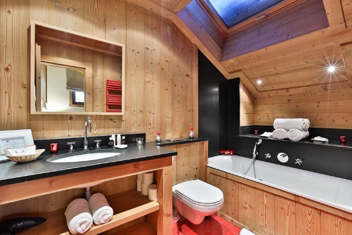 A cozy, wood-paneled bathroom with a modern sink, toilet, bathtub, and a skylight. Red accents and black elements add contrast.