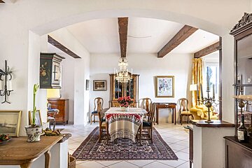 A charming, traditional dining room featuring wooden beams, a crystal chandelier, antique furnishings, and a vibrant Persian rug on white tiles.