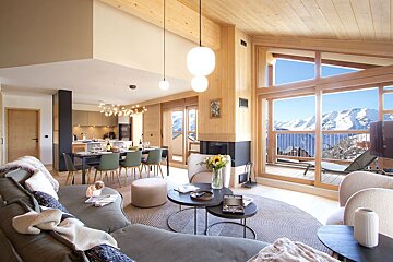 Modern, open-plan chalet living room with wood finishes and breathtaking snowy mountain views through large windows.