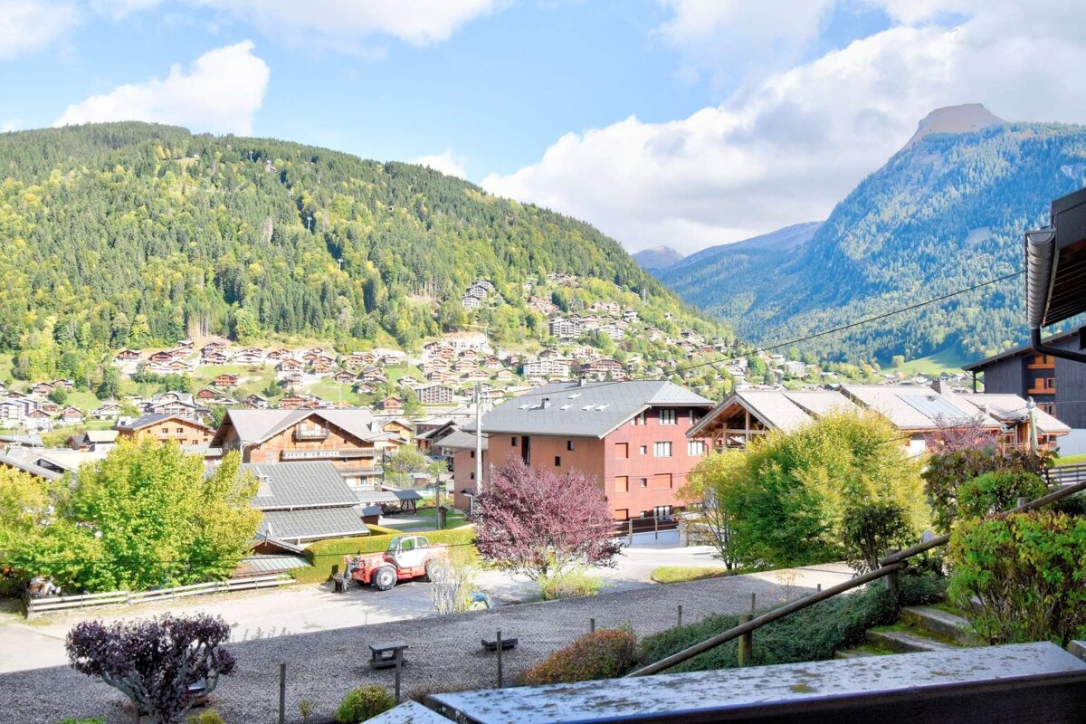 Charming mountain village with traditional houses nestled on lush, forested slopes under a bright blue sky, with a red tractor visible in the foreground.