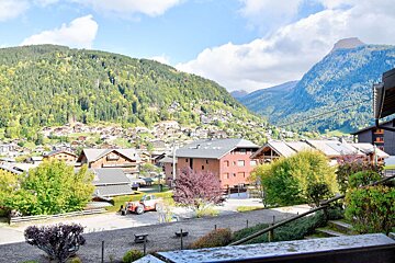 Charming mountain village with traditional houses nestled on lush, forested slopes under a bright blue sky, with a red tractor visible in the foreground.