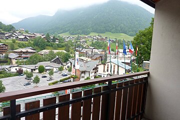 Balcony view of a green mountain village with houses, a parking lot, and international flags under a cloudy sky.