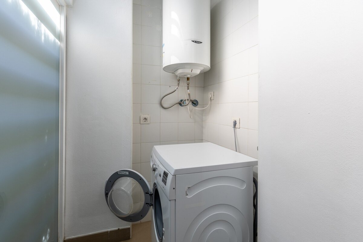A small utility room with white tiled walls, featuring a white washing machine with its door open and a white water heater mounted above it.