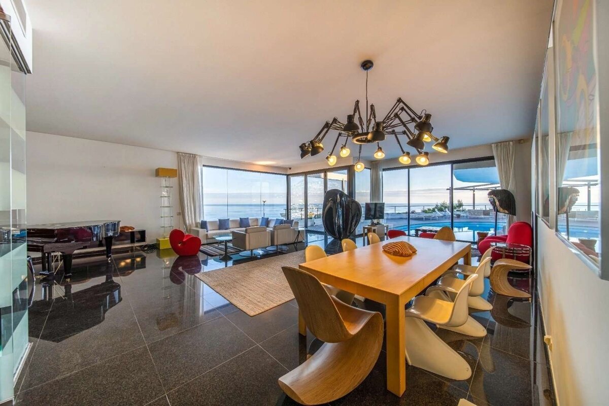 Luxurious, modern living/dining area with panoramic ocean and pool views. Features a grand piano, stylish furniture, and a unique spider-like chandelier.