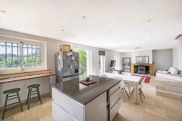 Bright, open-plan kitchen, dining, and living room with a kitchen island, dining table, fireplace, and scenic window view.