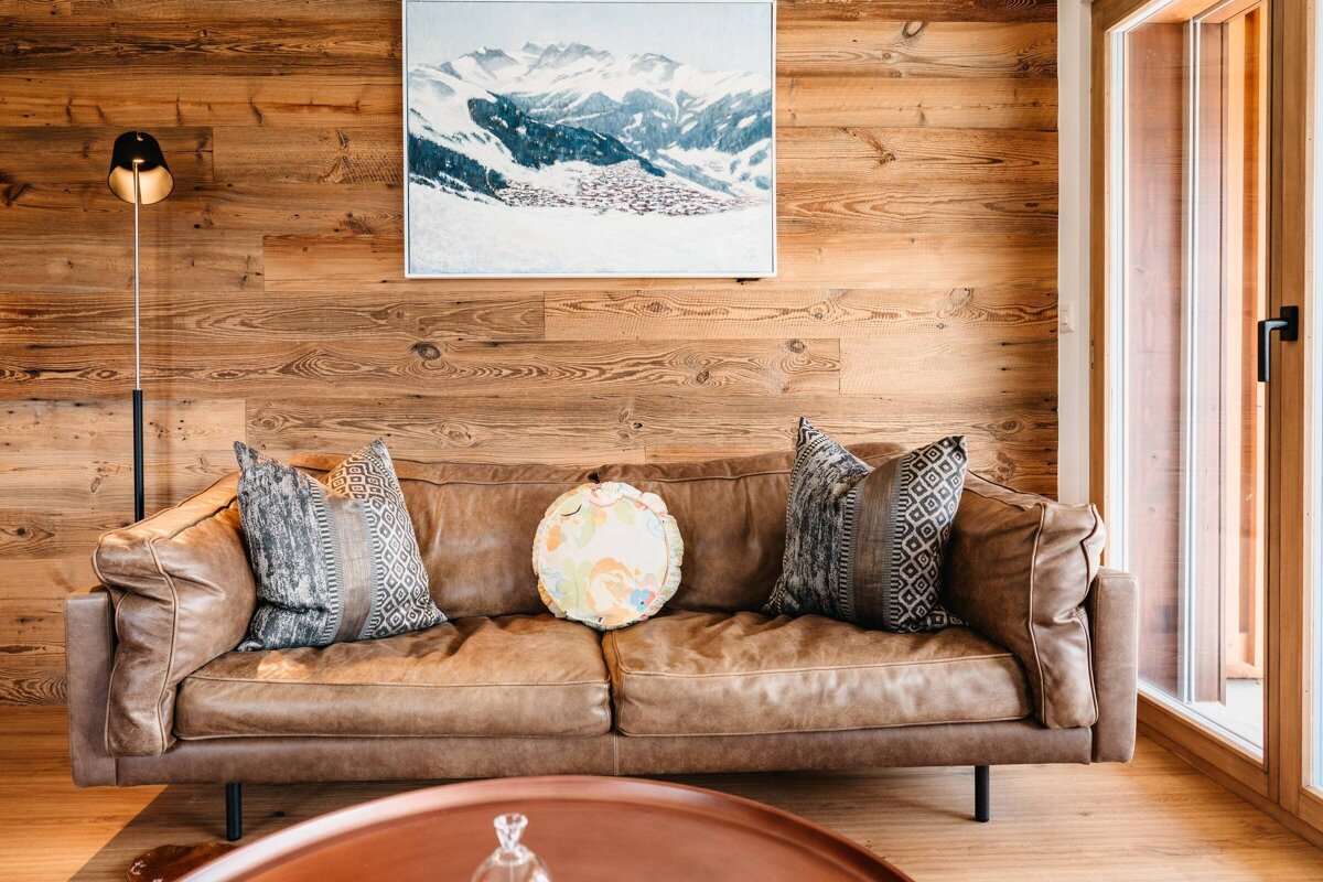 A brown leather sofa with patterned pillows against a rustic wood wall with a snowy mountain painting hanging above.