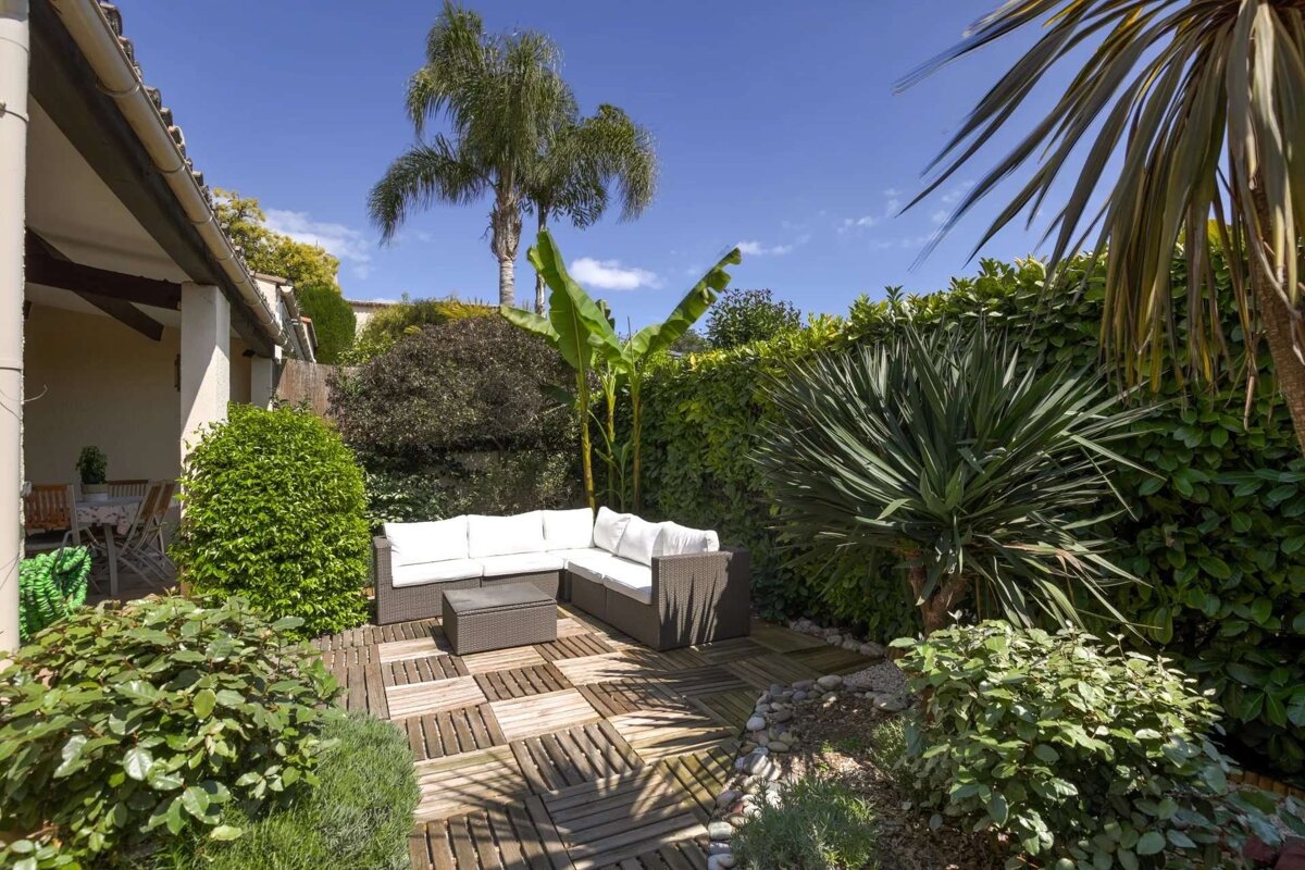 A sunny outdoor patio with an L-shaped wicker sofa, wooden deck, and abundant tropical plants, including palm trees and a banana plant, under a clear blue sky.