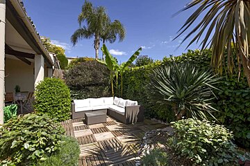 A sunny outdoor patio with an L-shaped wicker sofa, wooden deck, and abundant tropical plants, including palm trees and a banana plant, under a clear blue sky.