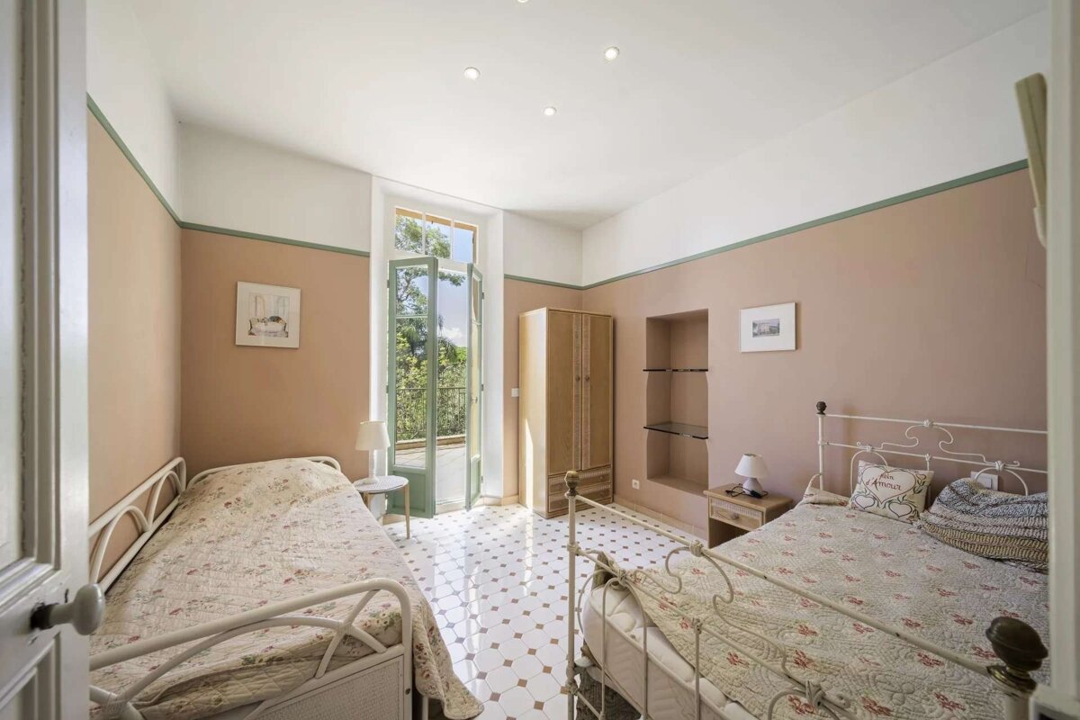 Sunny bedroom with two single beds, peach walls, patterned tiles, and open French doors leading to a green outdoor view.