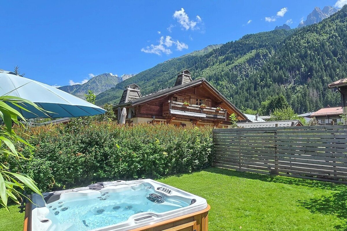 Scenic mountain view from a backyard hot tub, with a charming wooden chalet and lush forest under a clear blue sky.