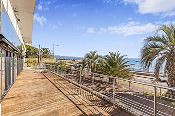 Spacious wooden deck with a metal railing, overlooking a sunny coastal road, blue sea, palm trees, and distant mountains under a clear sky.