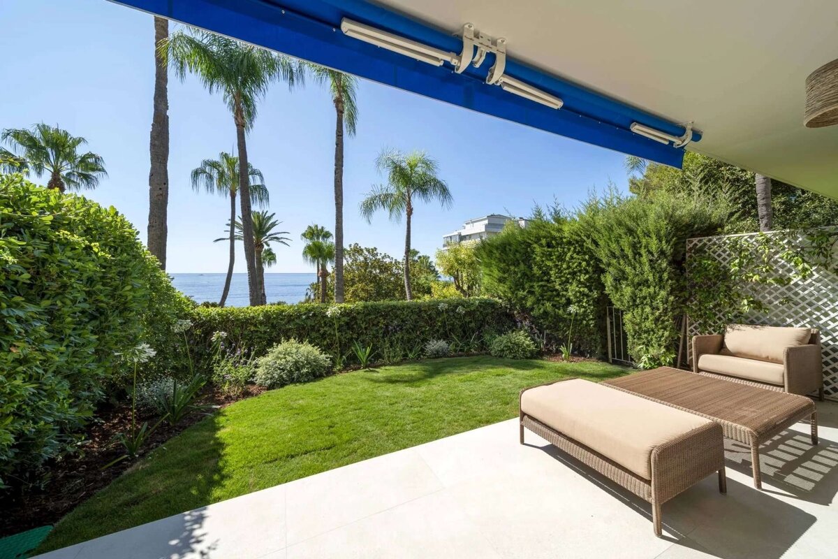 A patio with outdoor seating overlooks a vibrant green garden, palm trees, and the ocean, all beneath a bright blue awning.