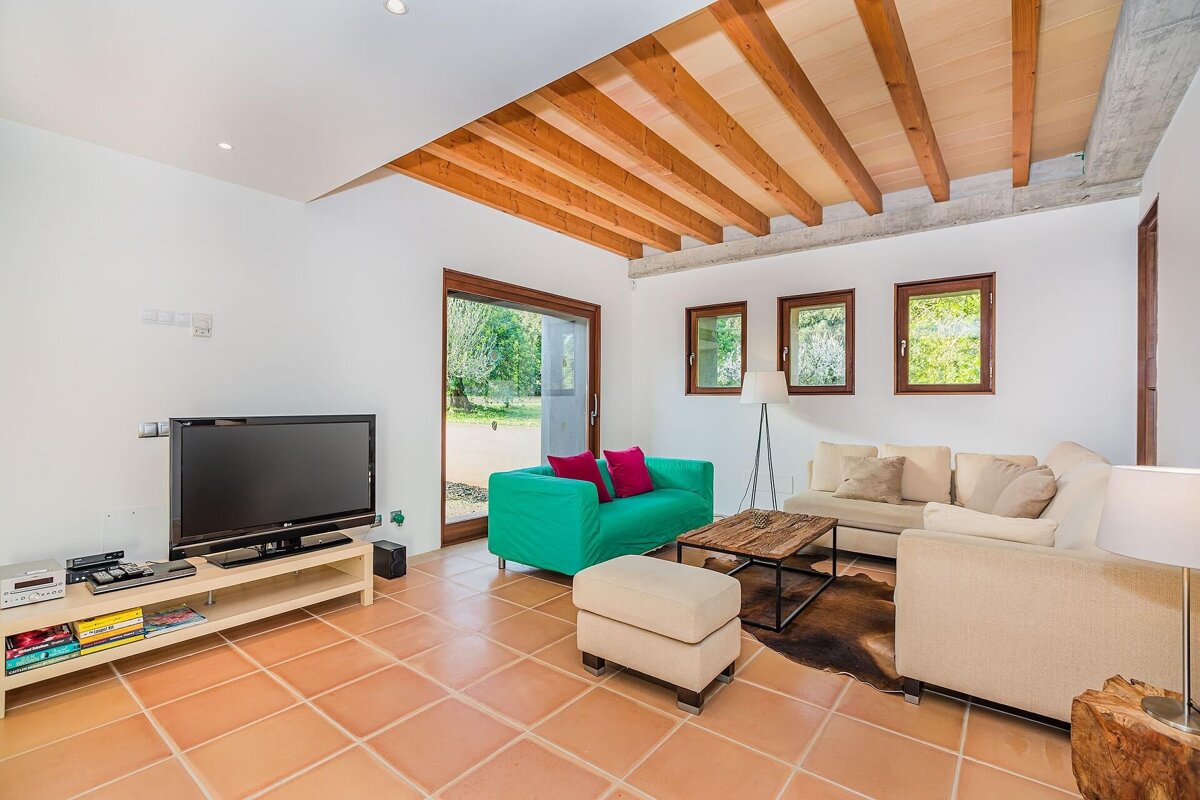 A bright, modern living room with rustic wooden ceiling beams, white walls, and terracotta floor. Features a green sofa, beige sectional, TV, and a large glass door to a garden.