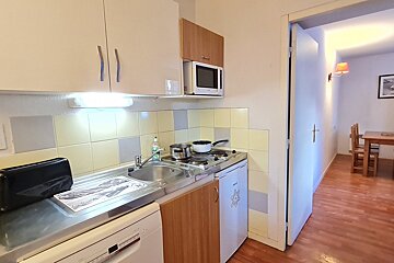 A compact kitchen area featuring a stove, sink, microwave, and small fridge, with light-colored cabinets and a tiled backsplash, leading to a dining space.