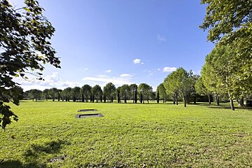 Sunny park with a vast green lawn, two embedded trampolines, and a distant line of trees against a blue sky.