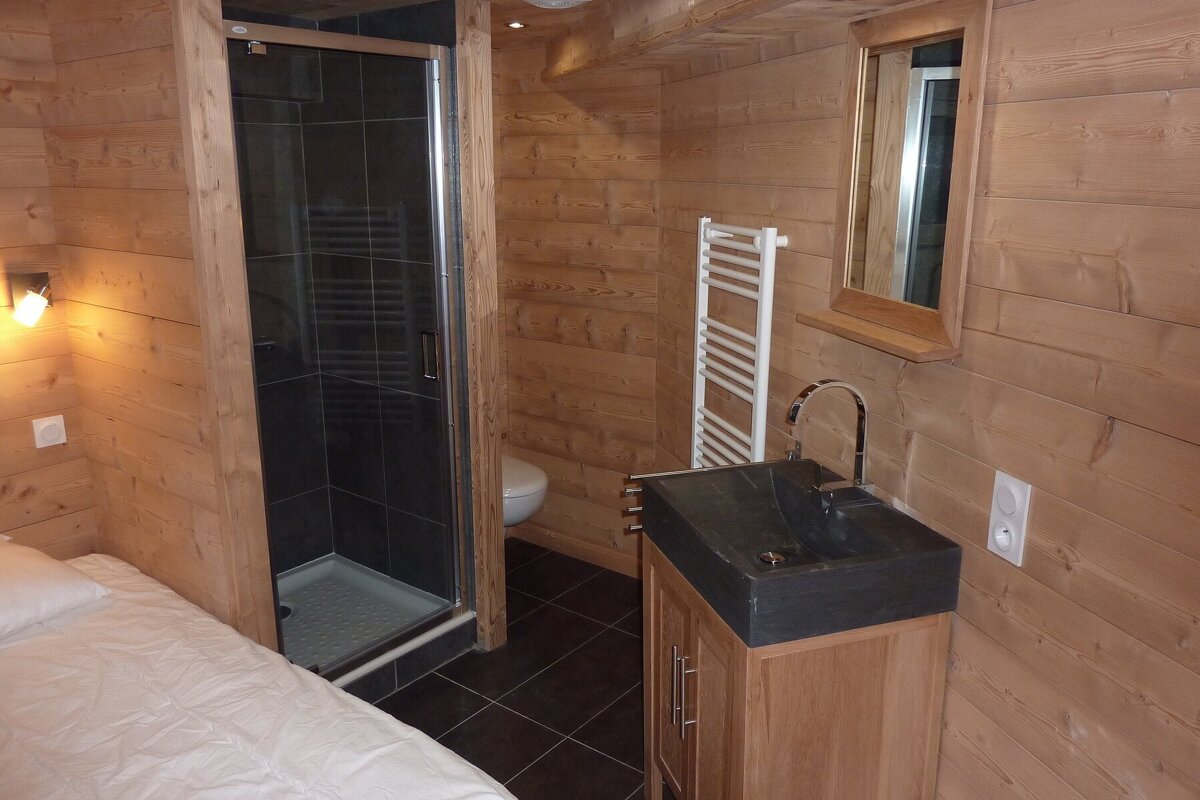 A compact, wood-paneled room with an integrated bathroom featuring a dark-tiled shower, toilet, and modern sink vanity. A bed is visible on the left.
