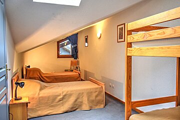 A cozy attic bedroom with a slanted ceiling, two single beds, and a wooden bunk bed. A small window and warm wall lighting are present.