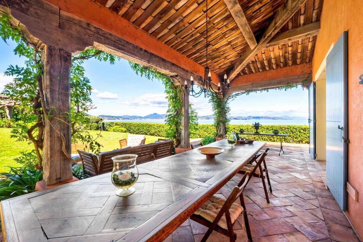 An outdoor patio with a long wooden dining table under a rustic wooden canopy, overlooking a green lawn and a body of water in the distance.