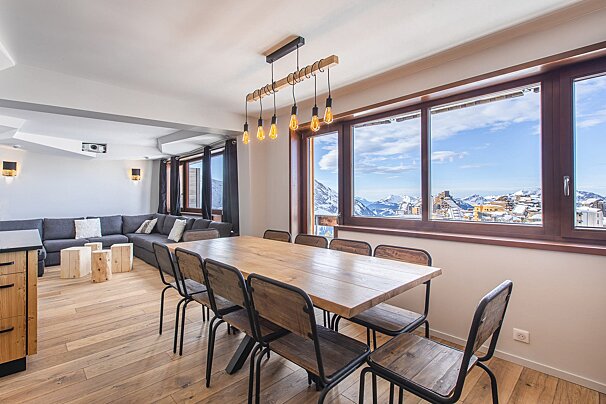 Bright modern dining room with a wooden table, industrial lighting, and large windows overlooking a snowy mountain ski resort.