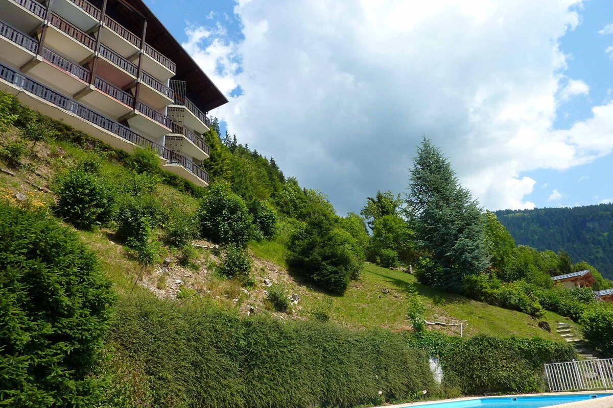 A multi-story building with balconies on a lush green hillside with trees, overlooking a pool under a partly cloudy sky.