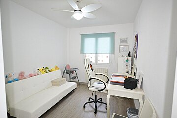 A bright white room with a sofa lined with plush toys, a desk with a gaming chair, and a window with a light blue blind.