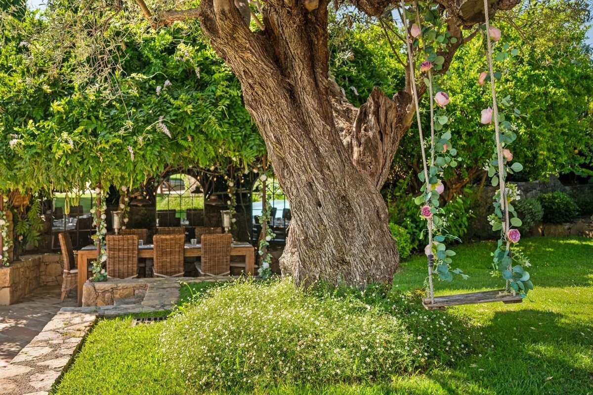 A lush garden scene features a large tree, an outdoor dining area under a leafy canopy, and a rustic swing adorned with flowers, creating a serene setting.