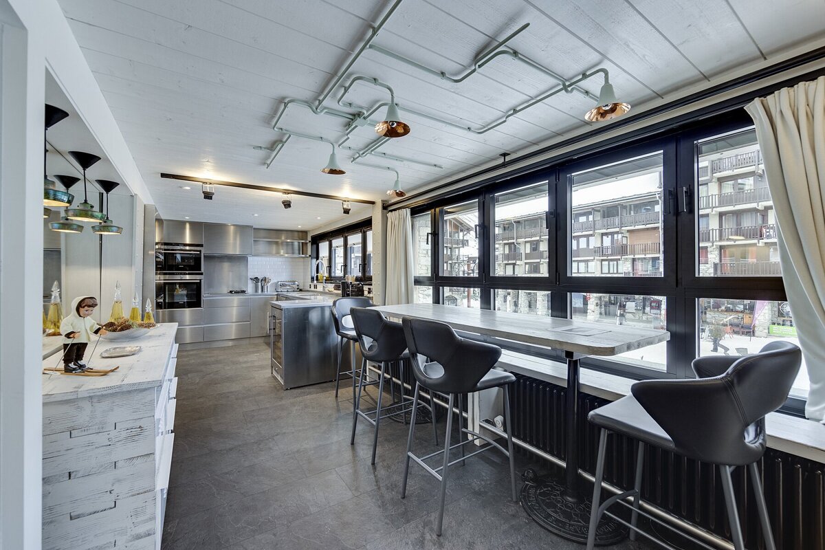 Sleek industrial kitchen with stainless steel appliances, exposed pipe lighting, and a long bar table by large windows viewing a resort town.
