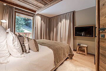 Cozy chalet bedroom with a large bed, balcony forest view, wood ceiling, light curtains, wall-mounted TV, and rustic details.