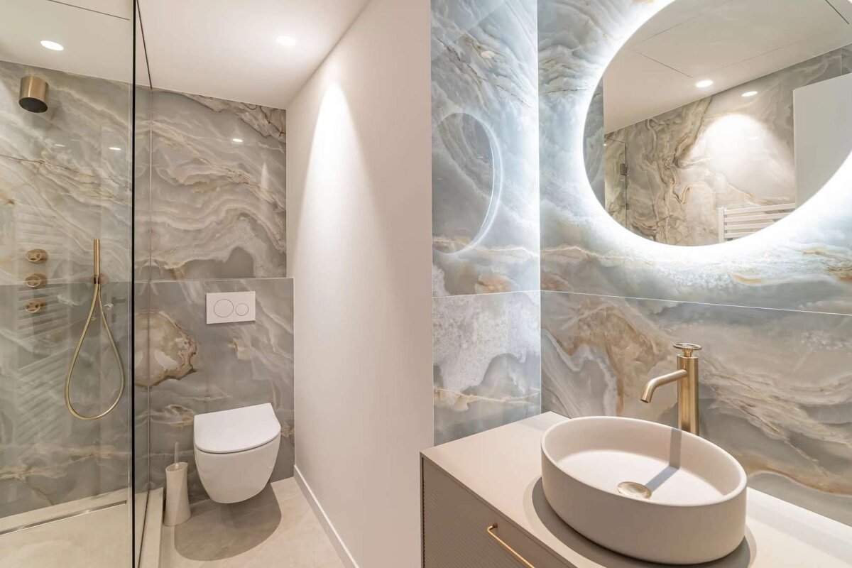 Chic bathroom with blue/grey marble tiles, gold fixtures, glass shower, wall-mounted toilet, and backlit mirror above vanity.