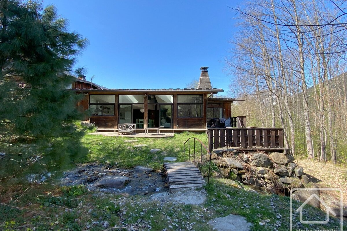 A modern wooden house with large windows overlooks a flowing stream and small bridge, set amidst trees under a clear blue sky.