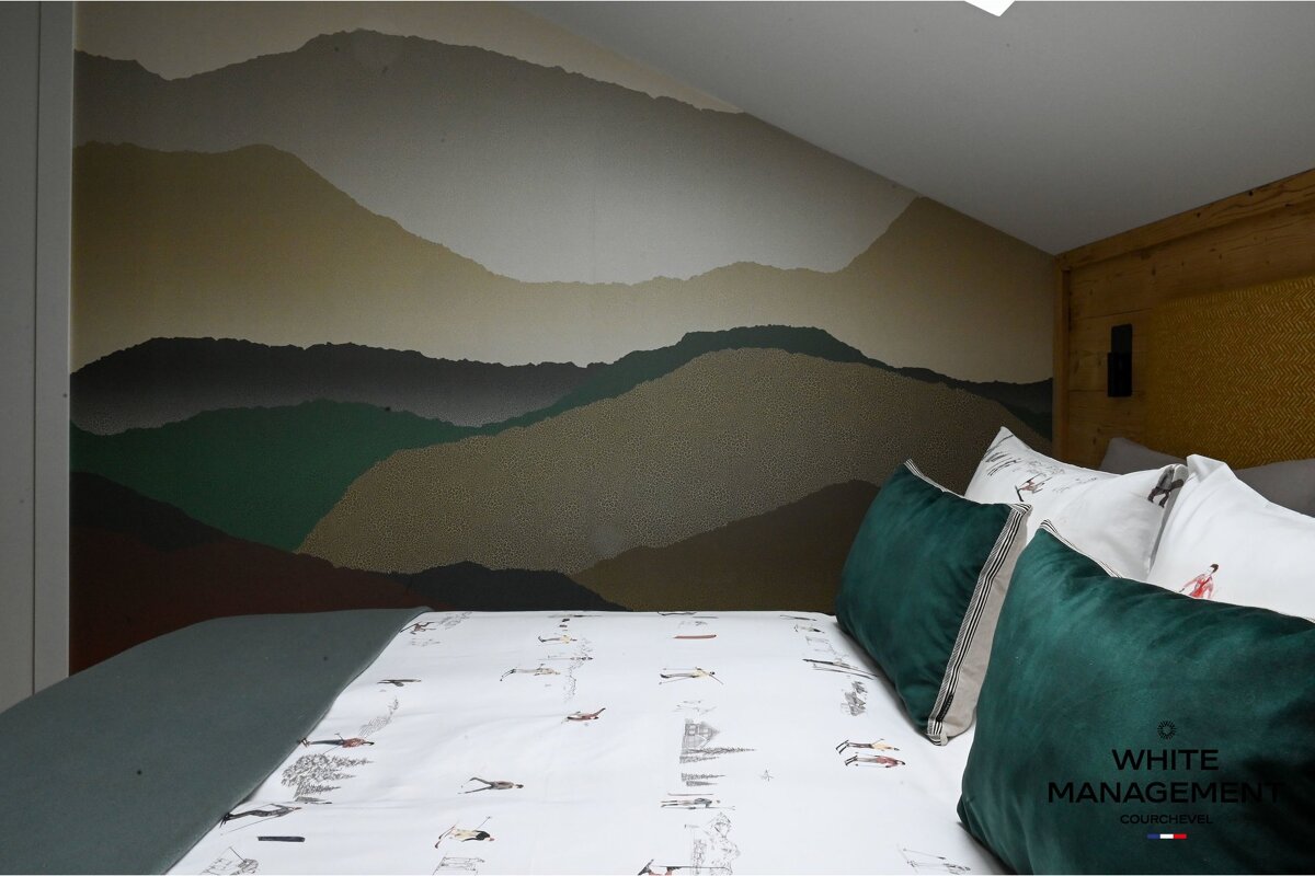 A cozy bedroom with a mountain landscape mural behind a bed featuring ski-themed bedding, dark green pillows, and a textured wooden headboard.