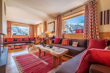 A warm chalet living room with dark sofas, red accents, wooden furniture, and panoramic windows overlooking snow-covered mountains.