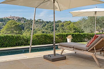 A patio with umbrellas, a pool, and a lounge chair overlooks a lush, hilly landscape with a distant town under a clear sky.