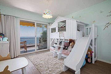 A bright children's bedroom features a white playhouse bunk bed with a slide, a fluffy rug, and large glass doors opening to a balcony with an ocean view.