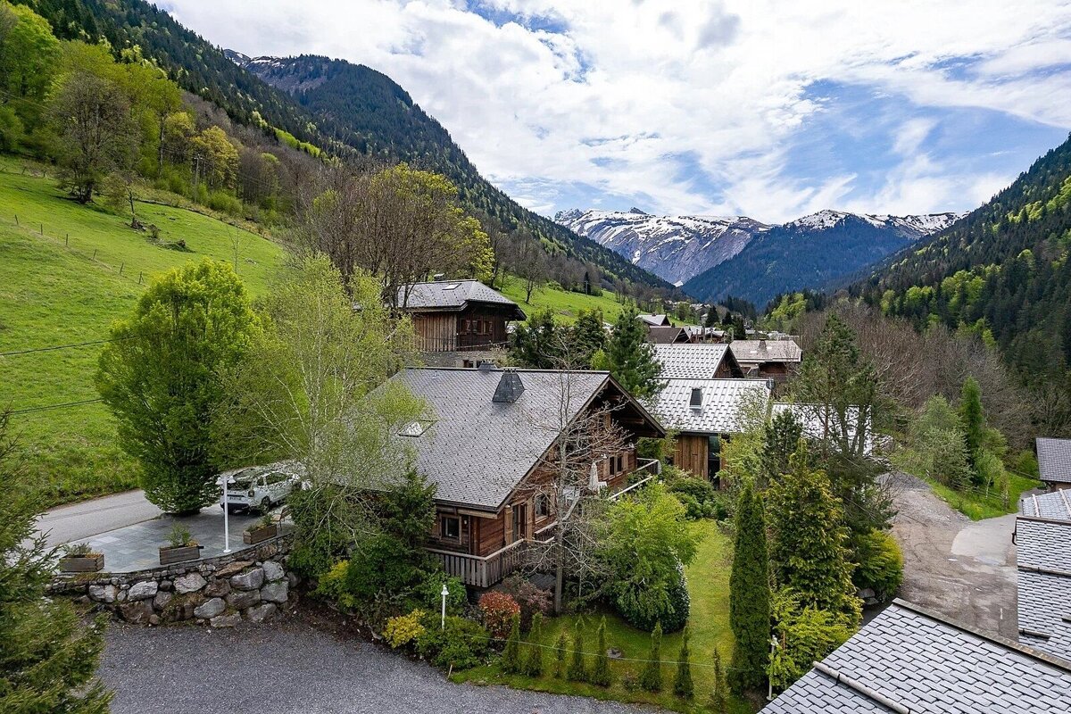 An aerial view of a charming alpine village with wooden chalets amidst lush green mountains and distant snow-capped peaks under a partly cloudy sky.