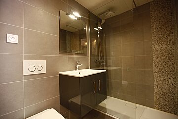 Modern tiled bathroom with gray walls, a vanity, mirror, and a glass-enclosed shower. A toilet and power outlet are also visible.
